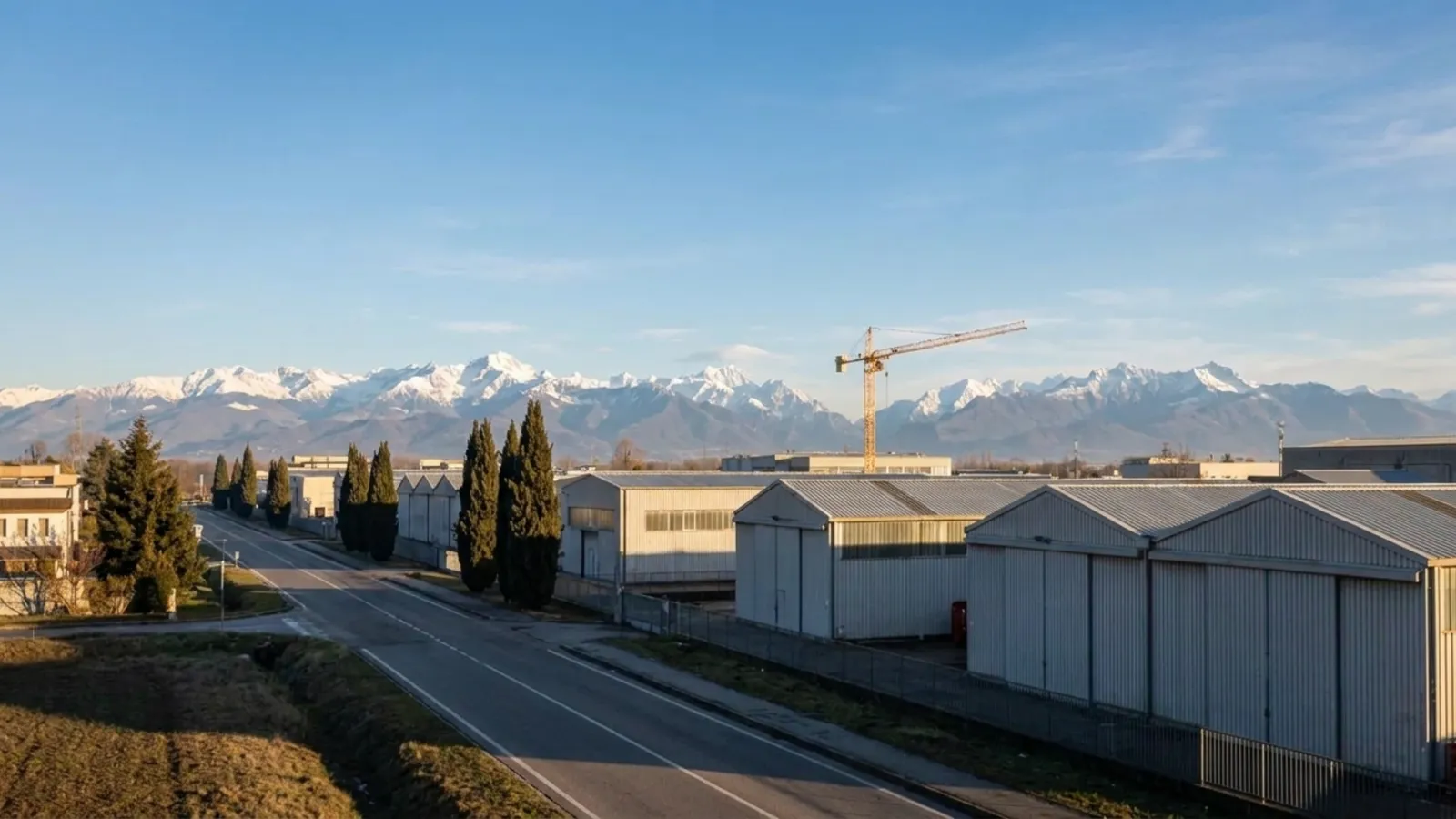 Northern Italian industrial plain at dusk with an Alpine silhouette behind and one orange tower crane